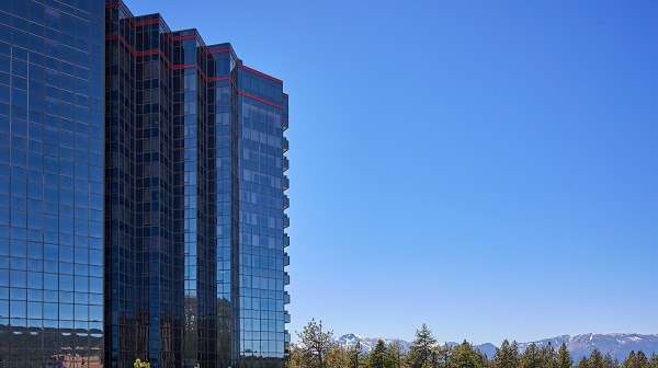 A tall glass building with red trim stands against a clear blue sky, with trees and mountains in the background.